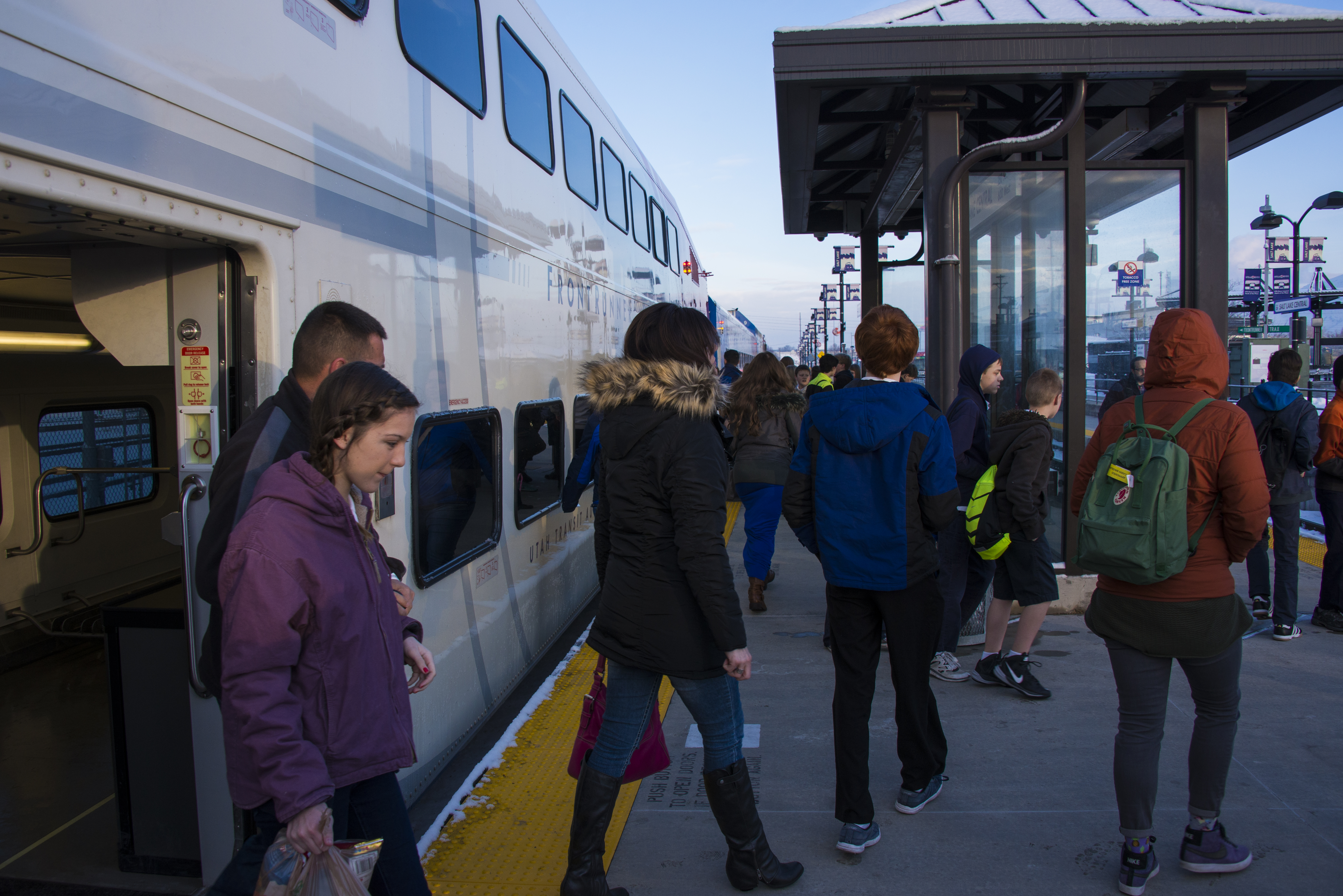 A group of about a dozen riders wearing winter coats exit from a FrontRunner train car onto a station platform.